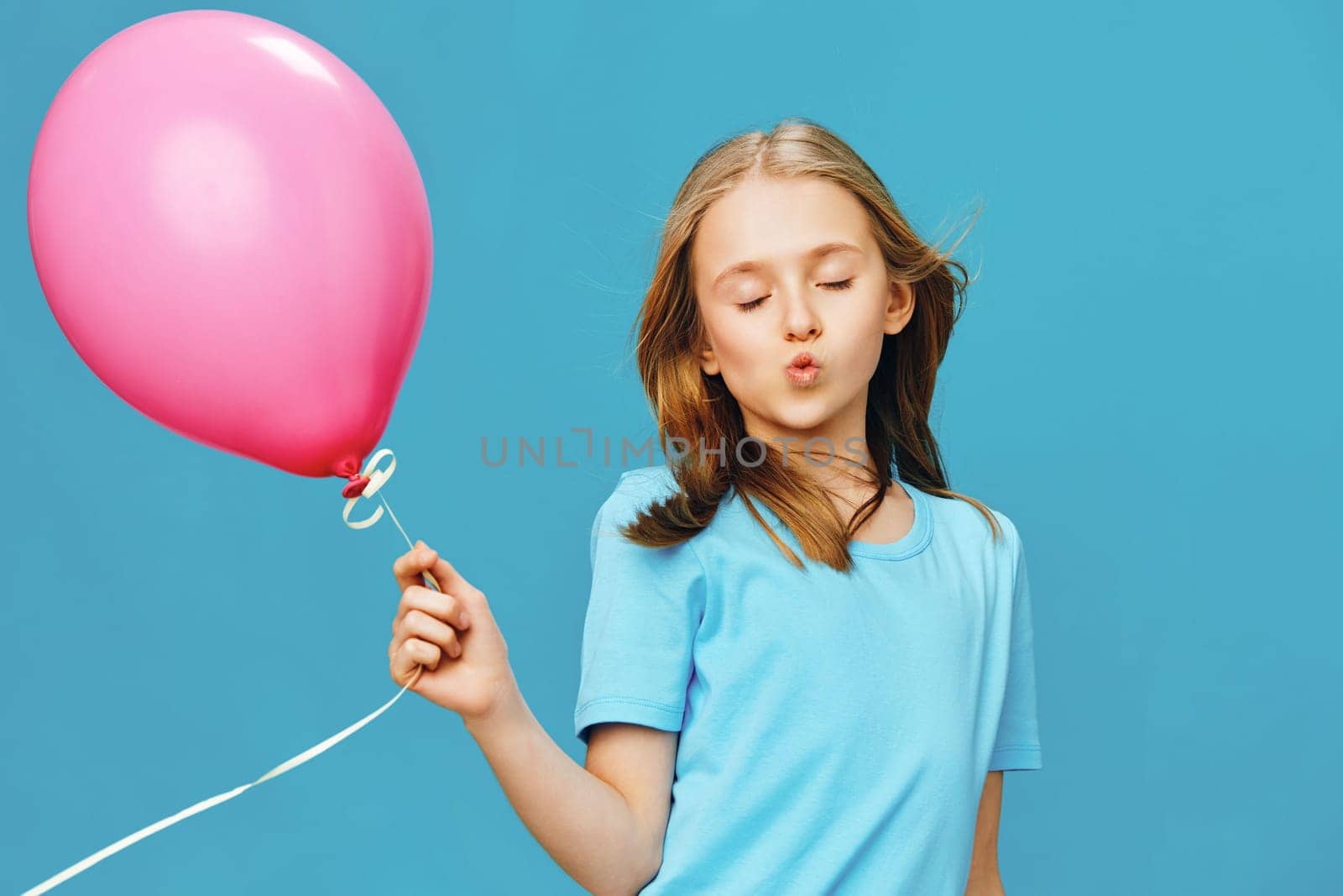 Joyful girl with long hair holding a pink balloon, wearing a blue shirt, enjoying a carefree moment against a vivid blue background, radiating happiness and innocence