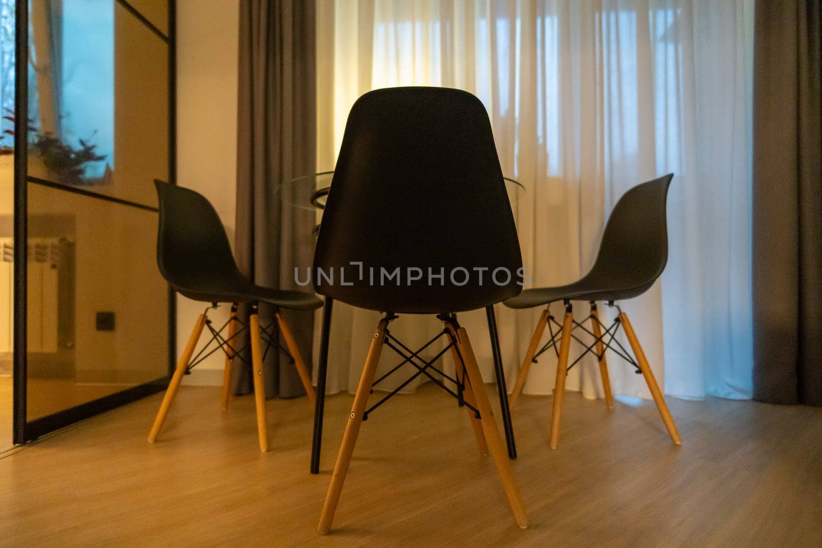 Three black Eames-style chairs surround a small dining table in a brightly lit room with sheer curtains and a mirrored partition.