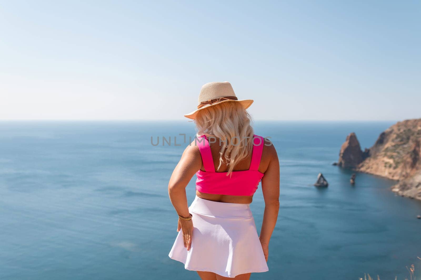 A woman in a pink top and white skirt stands on a cliff overlooking the ocean. The scene is serene and peaceful, with the woman taking in the beauty of the view