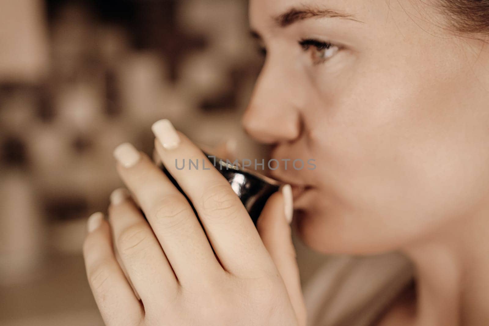 Close-up profile of a woman with light skin and manicured nails delicately sipping from a small, dark-colored cup.