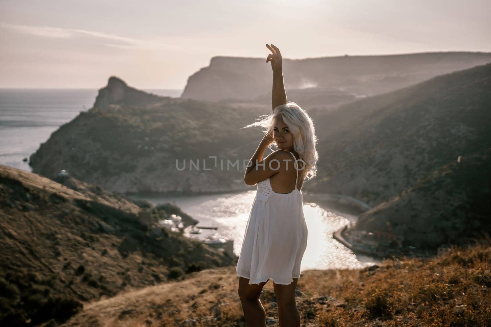 A woman stands on a hill overlooking a body of water. She is wearing a white dress and she is enjoying the view