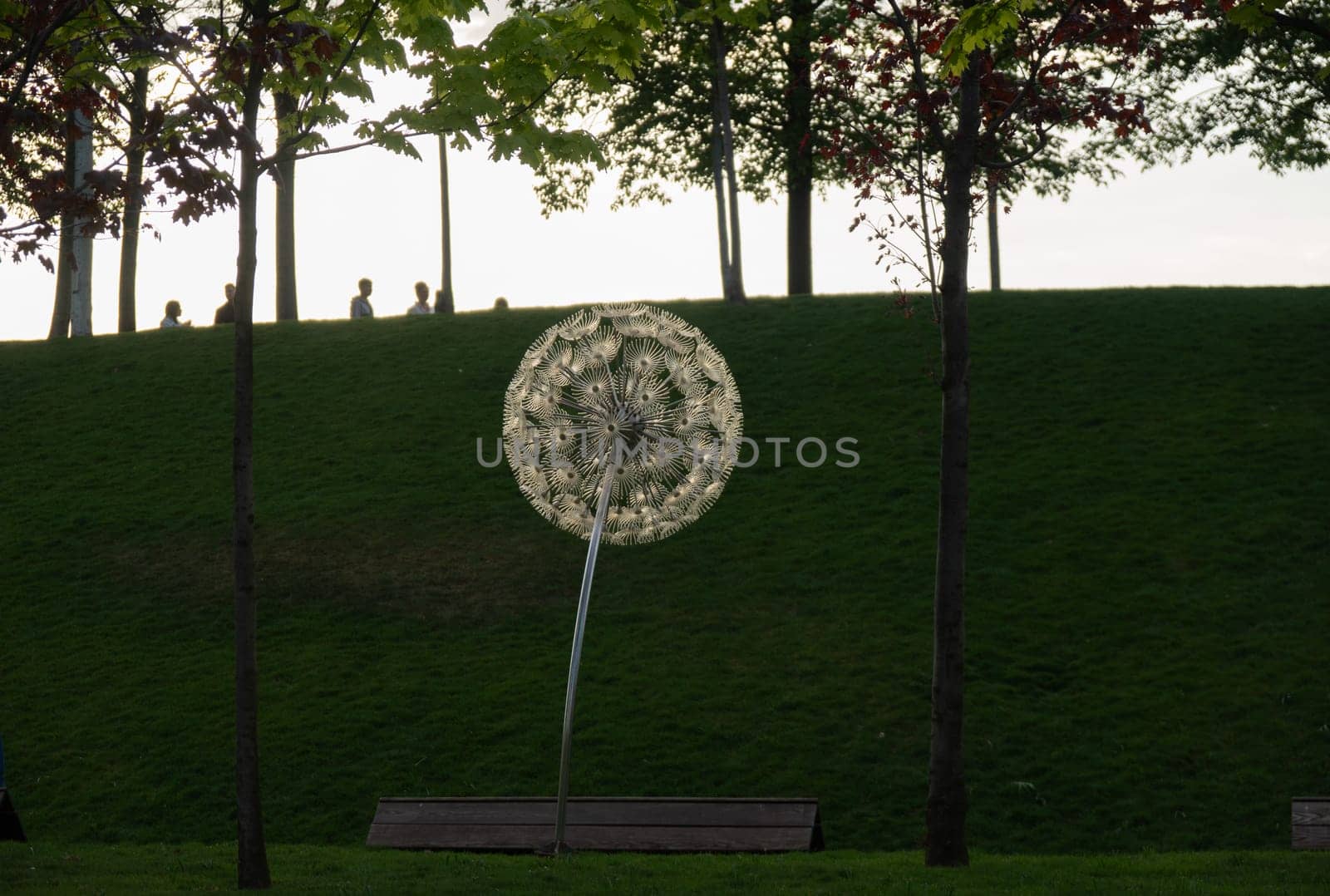 A large, white, circular sculpture resembling a dandelion stands on a grassy hill. People are visible in the distance on the hill. The sculpture is surrounded by trees and greenery.