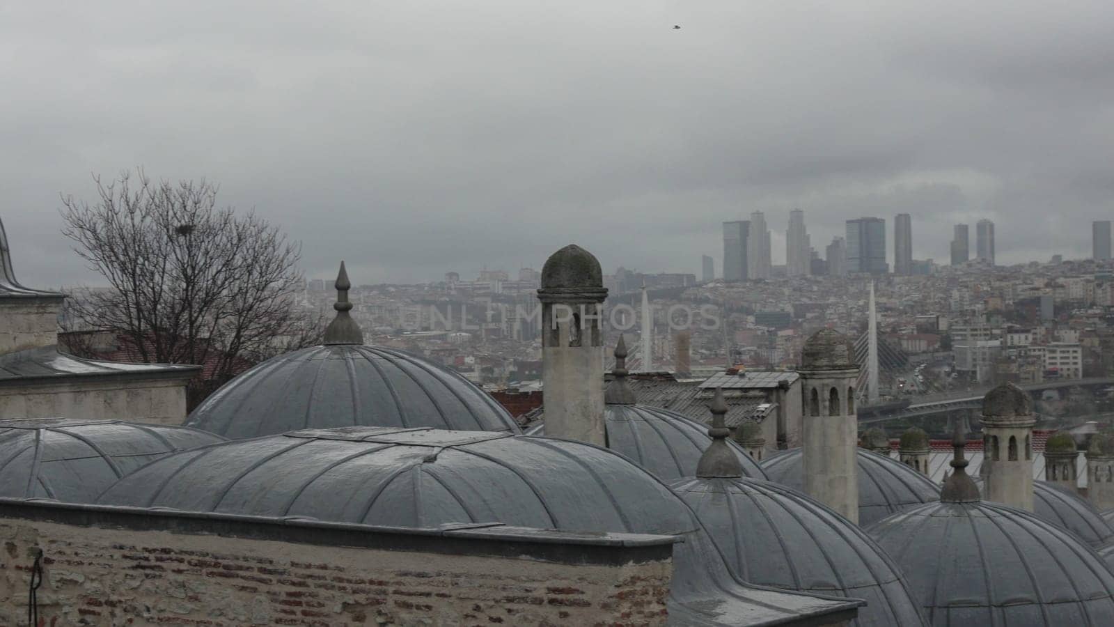 Overcast Istanbul skyline view from a rooftop. Grey domes, chimneys, and a sprawling cityscape beneath a cloudy sky. Modern skyscrapers visible in the distance.
