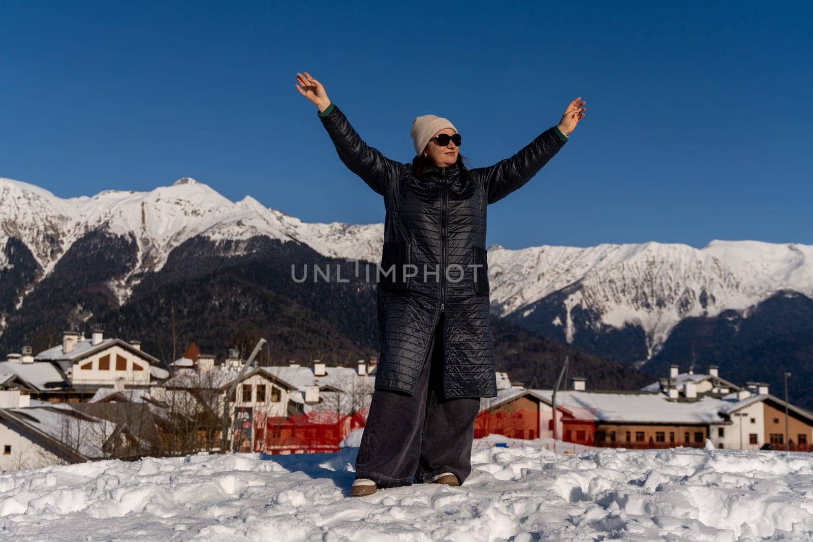 Woman in long coat joyfully raises arms, overlooking snow-covered village nestled in majestic, snow-capped mountains under a vibrant blue sky.
