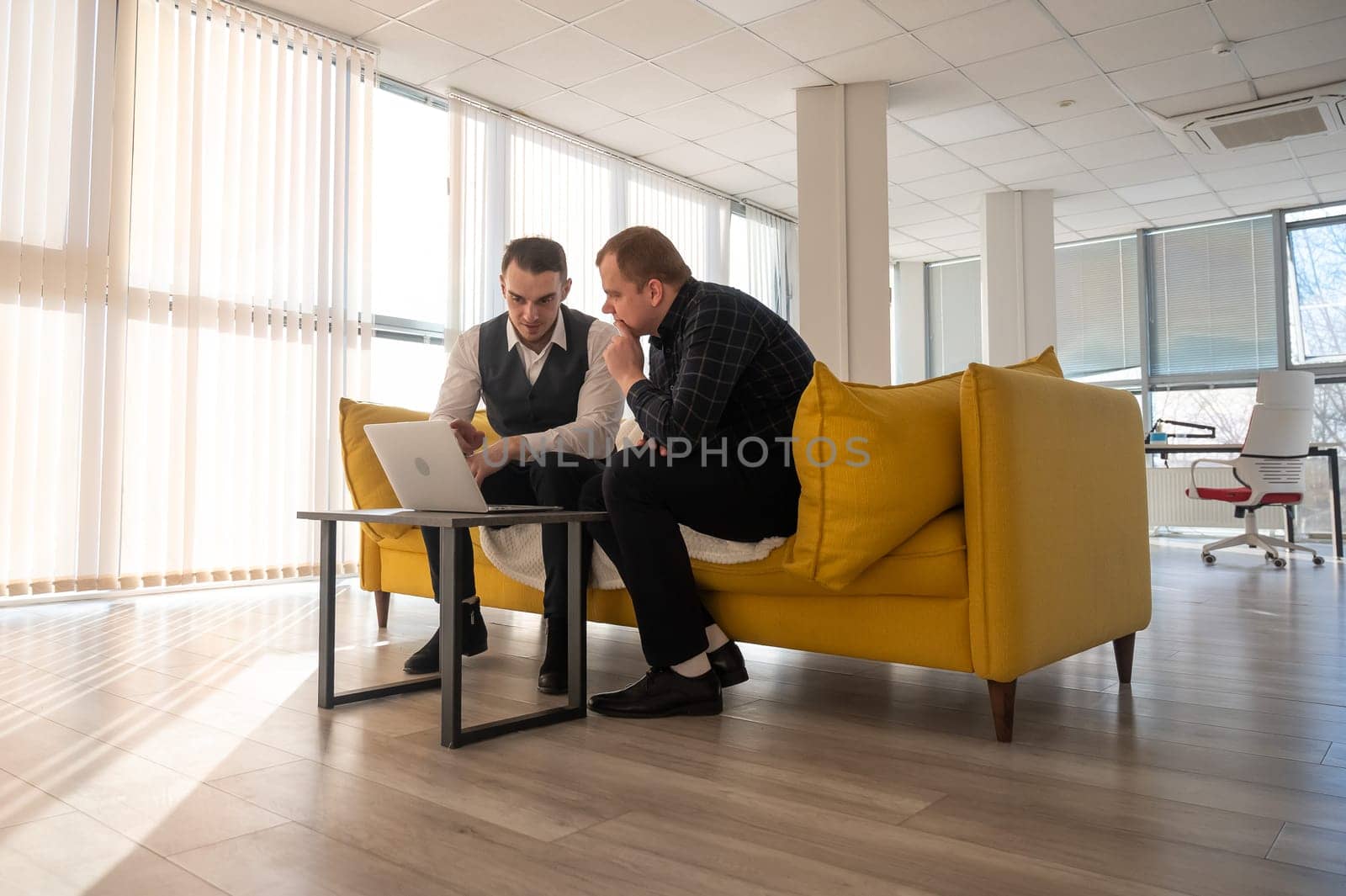 Two Caucasian men discussing work on a laptop while sitting on a sofa. Modern office