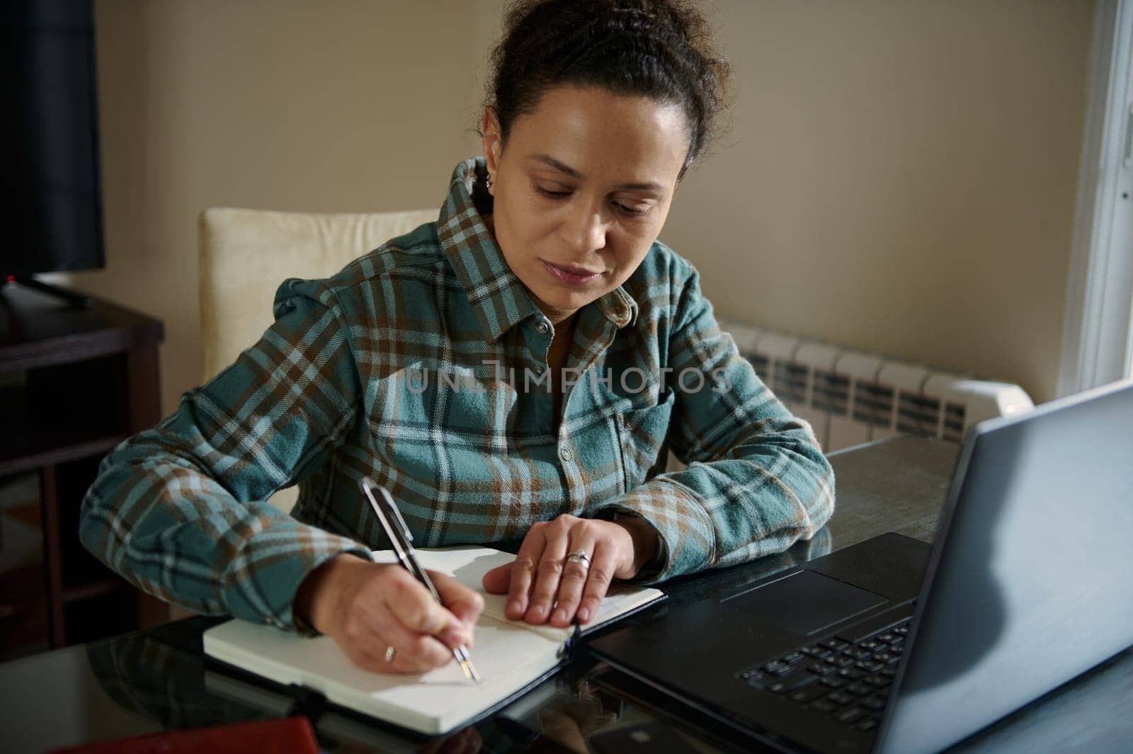 A woman focuses on her tasks, using her laptop and writing notes, seated in a comfortable home workspace. The image reflects a dedicated and productive atmosphere.