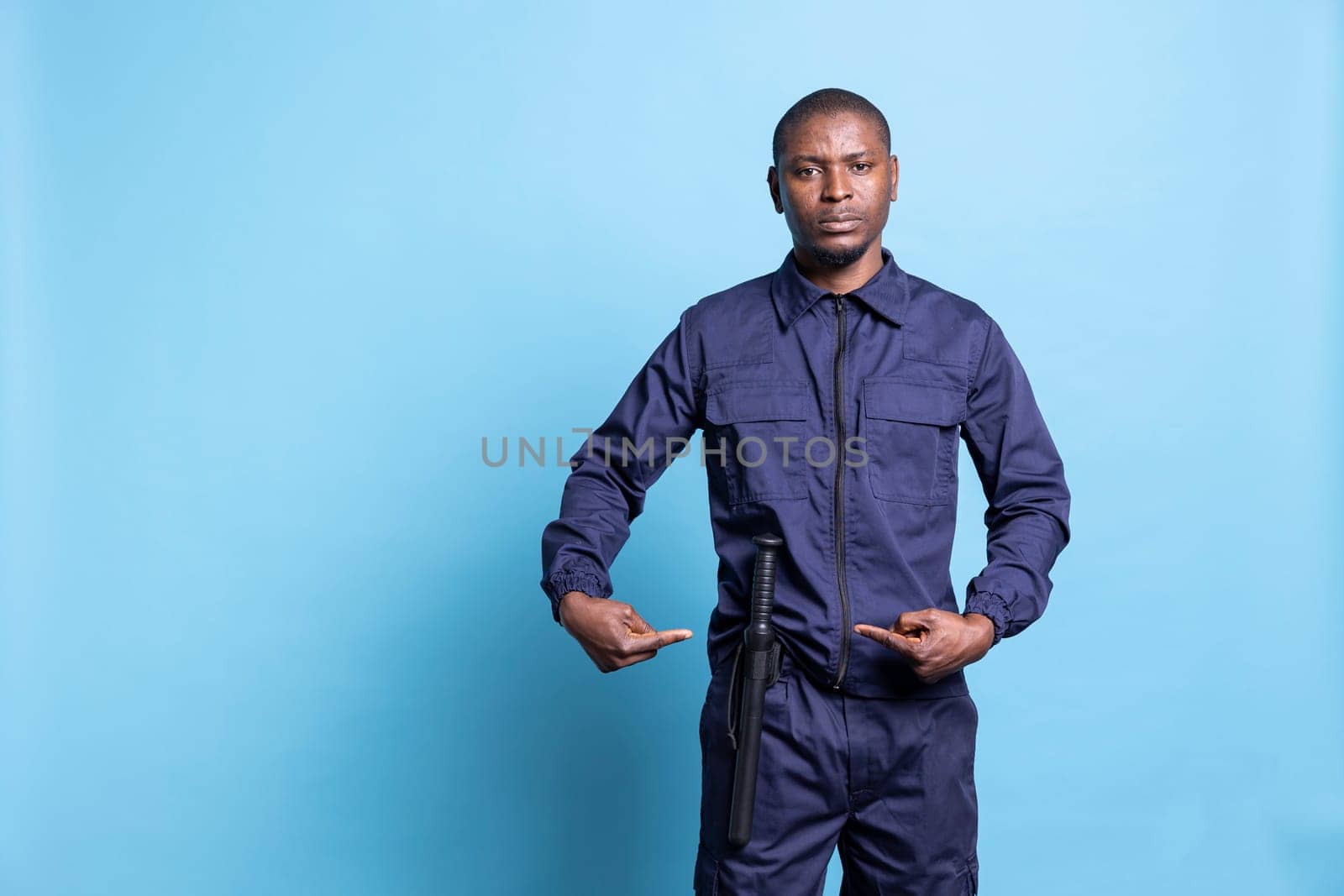 Armed security officer presenting his baton weapon for own safety in studio, pointing at his police bat to ensure order and protection. Male trained personnel in uniform against blue background.