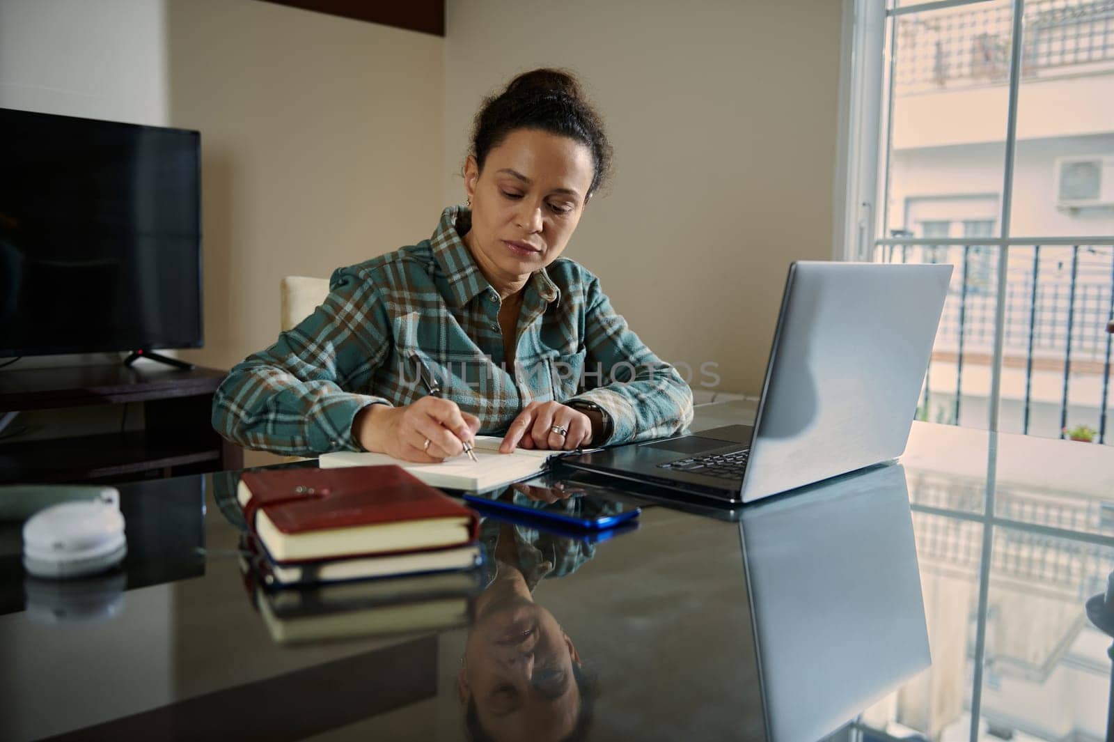 A focused woman works remotely, writing notes while engaging with her laptop in a home setting. The image exudes productivity, determination, and the blend of traditional and digital tools.