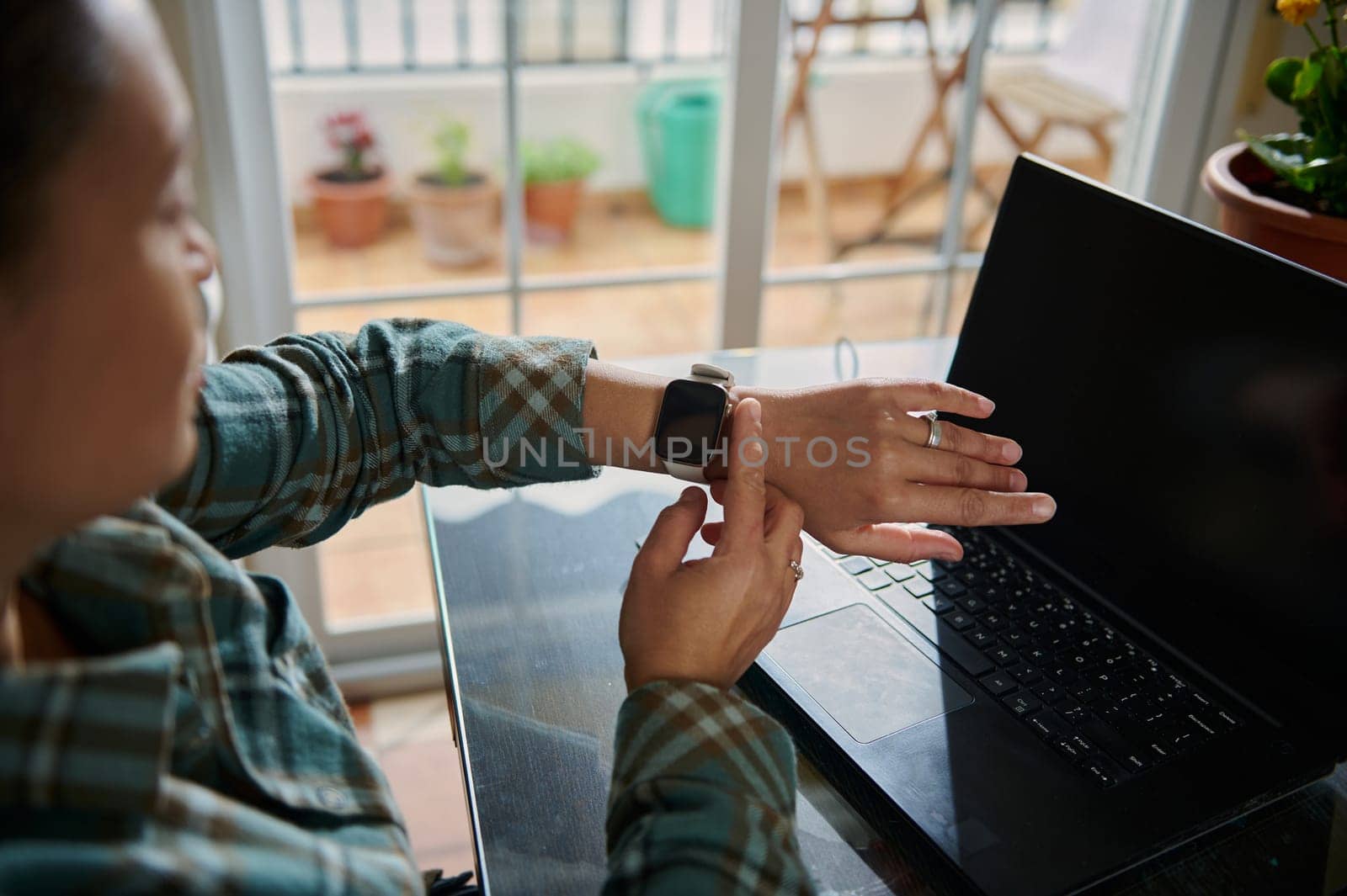 A person interacts with their smartwatch while working on a desk with a laptop in a cozy home environment, illustrating modern technology and productivity.