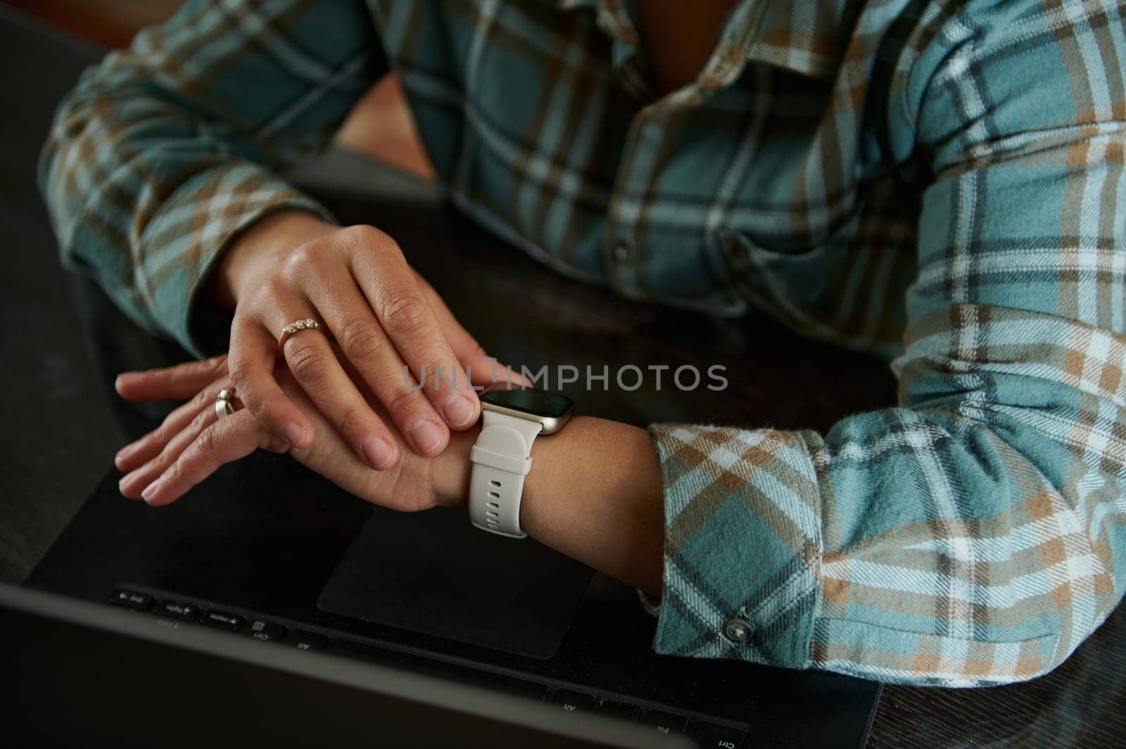 A close-up of hands resting on a smartwatch over a laptop. The individual wears a flannel shirt, emphasizing a relaxed home office setting and symbolizing modern technology usage.