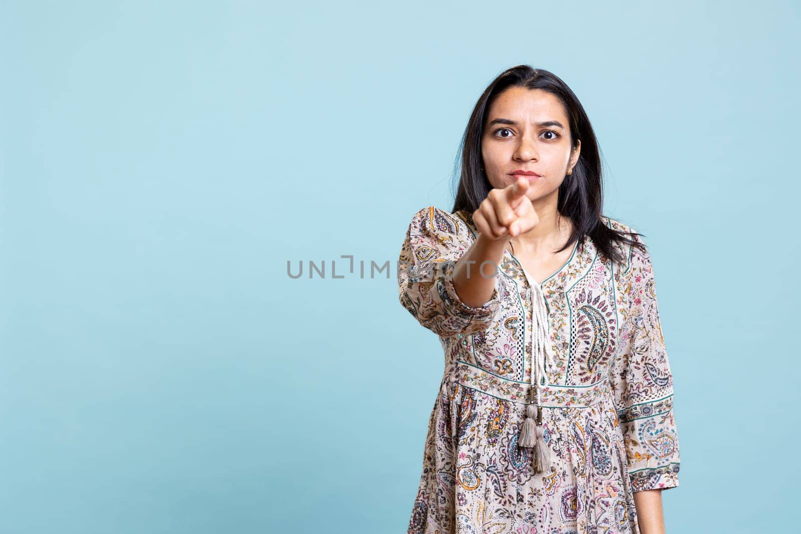 Indian serious woman pointing her index finger at the camera and looking forward, choosing someone and acting calm against blue background. Young person satisfied making a choice.