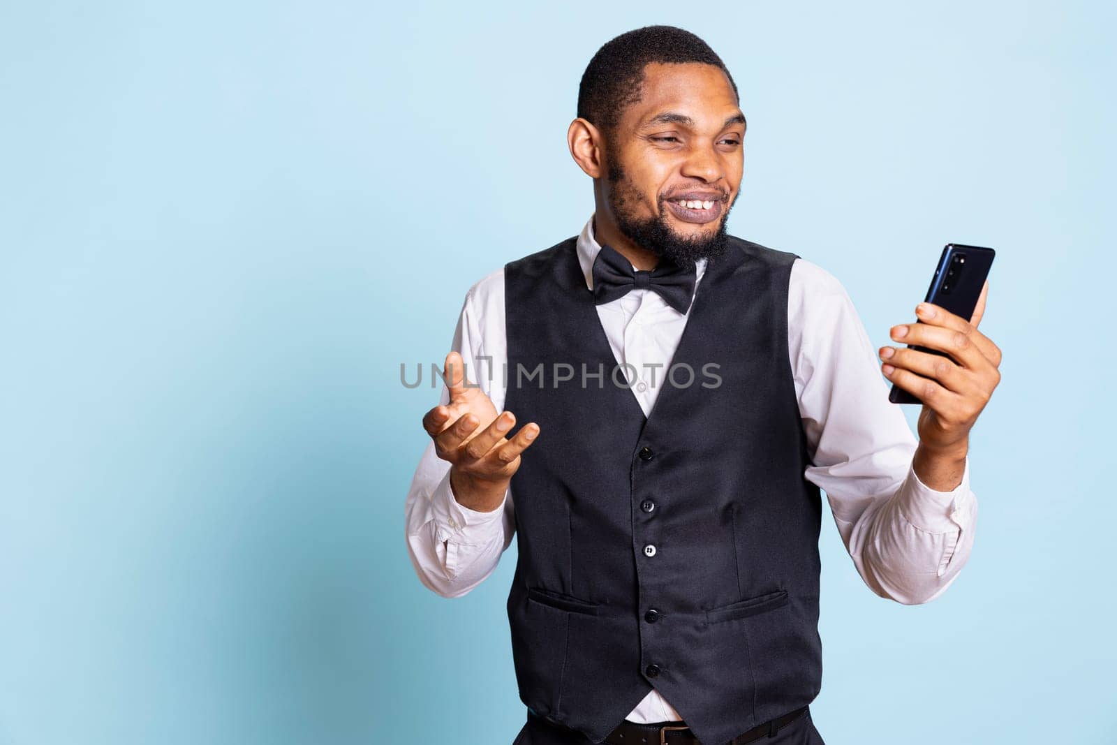 Doorman with suit and tie answering online videocall on phone, discussing with clients or guests against blue background. Bellboy staff on videoconference connection in studio.