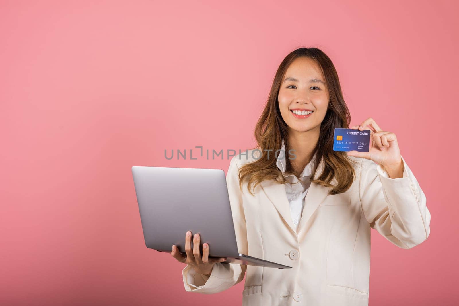 Portrait of Asian beautiful young woman customer showing credit card for online payment shopping transactions cashback loan on laptop studio shot isolated on pink background