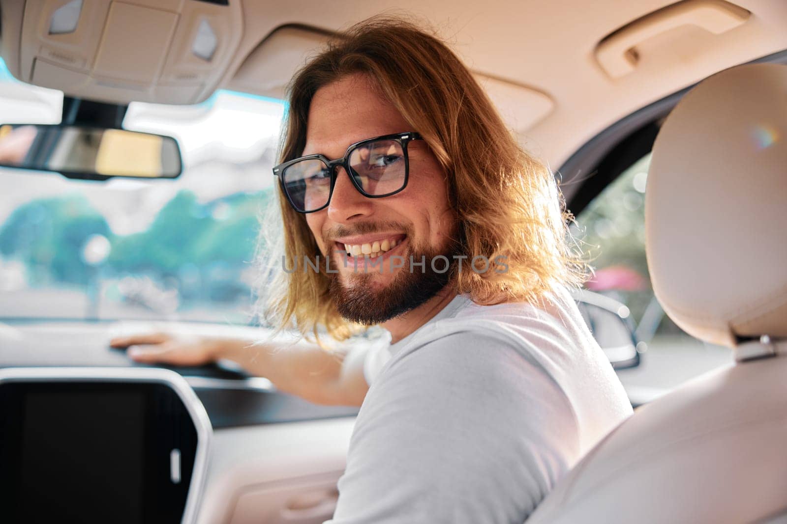 Happy young man with long hair and glasses in a car, looking back with a smile, creating a warm atmosphere