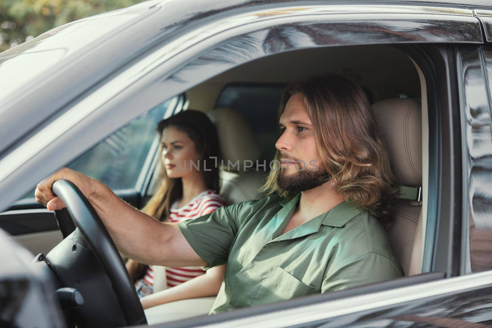 A man and woman sit in a car, the man driving, focused on the road ahead, while the woman gazes away, lost in thought
