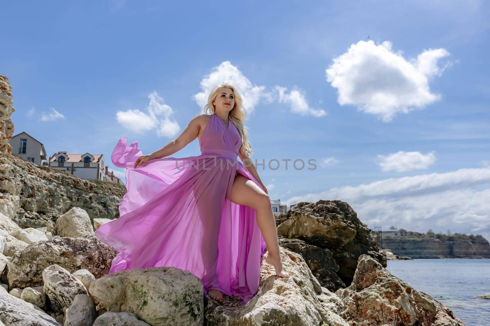 A blonde woman in a flowing pink dress poses on rocky coast, with a blue sky and ocean background.