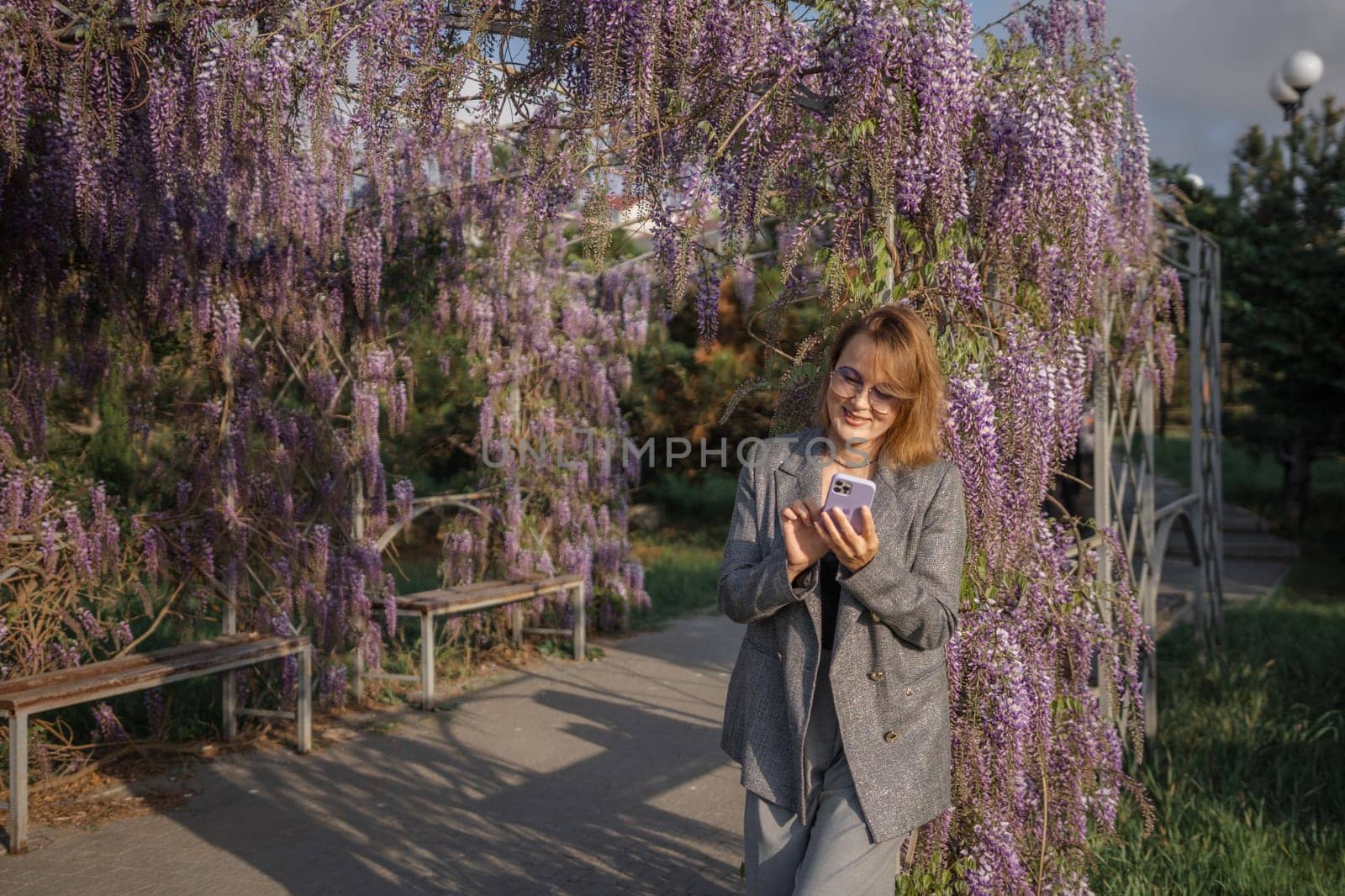 A woman in a grey blazer walks through a lush, purple wisteria trellis, holding a book. She smiles at the camera. A bench sits beneath the wisteria.