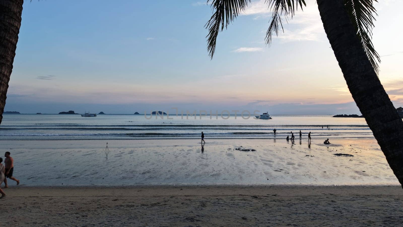 Tourists are walking and playing on a beautiful tropical beach at sunset, with palm trees framing the scene and a yacht sailing in the distance