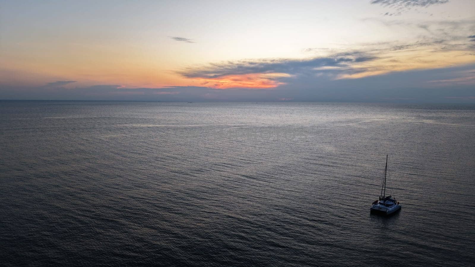 Catamaran sailing peacefully in a calm sea as the sun sets, casting a warm glow on the water and clouds, creating a serene and tranquil atmosphere