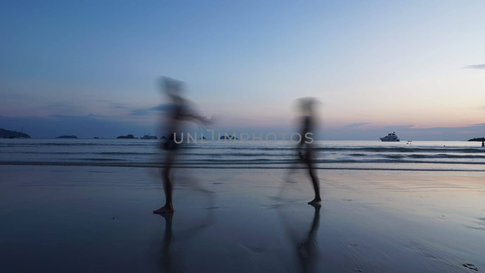 Two tourists are running along the beach at dusk, their figures blurred by motion, with a boat visible in the distance and the sky reflecting in the wet sand