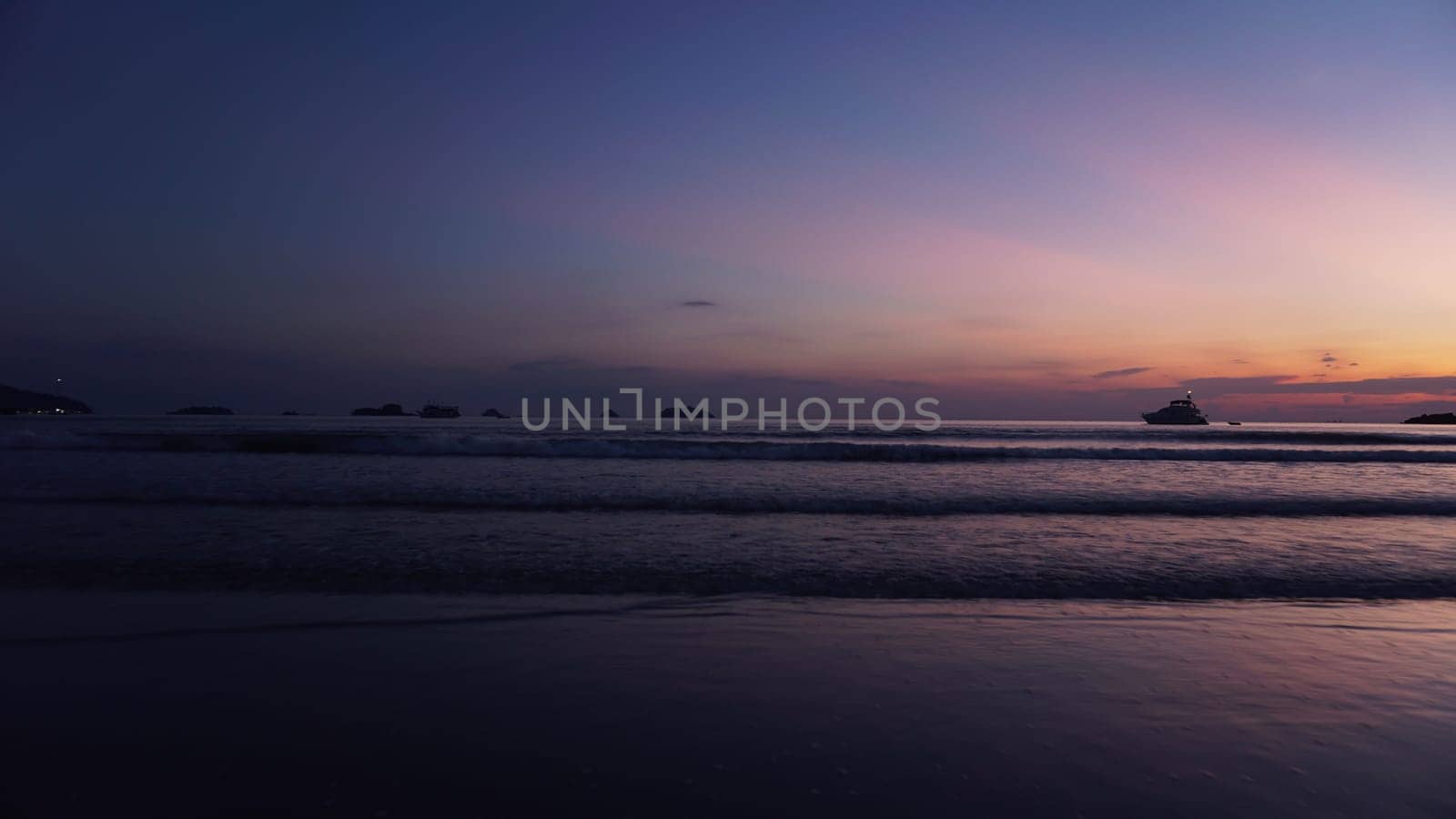 Gentle waves lapping at the shore during sunset, creating a tranquil coastal scene with a solitary boat silhouetted against a vibrant, colorful sky filled with hues of orange, pink, and purple