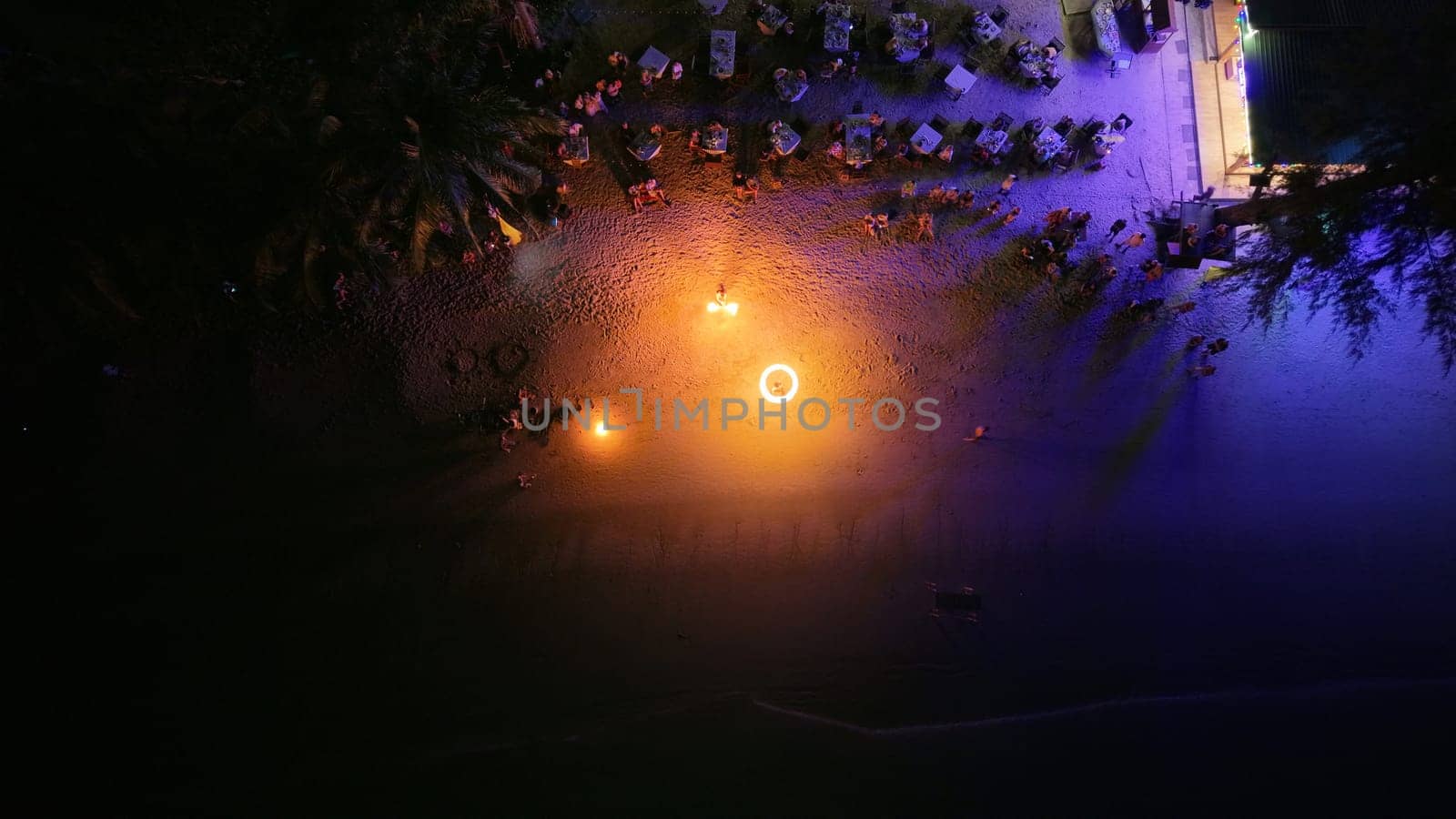 Aerial view capturing a vibrant beach resort at night, with a mesmerizing fire show lighting up the sandy shore as tourists gather along the coastline, enjoying the festive ambiance