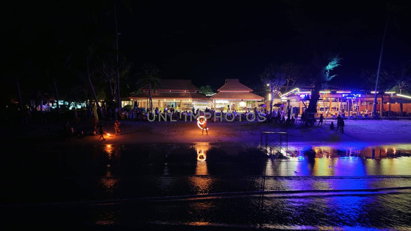 Tourists are relaxing and enjoying a fire show on a beach at night, with restaurants and palm trees in the background, creating a vibrant and tropical atmosphere