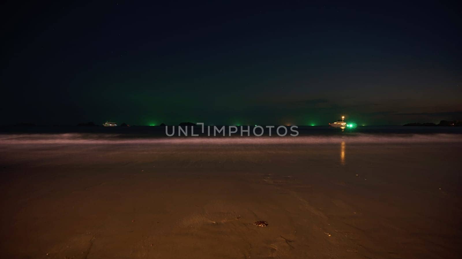 Long exposure capturing fishing boats glowing with green light on a tranquil sea, with a sandy beach in the foreground under the night sky, creating a serene and picturesque scene