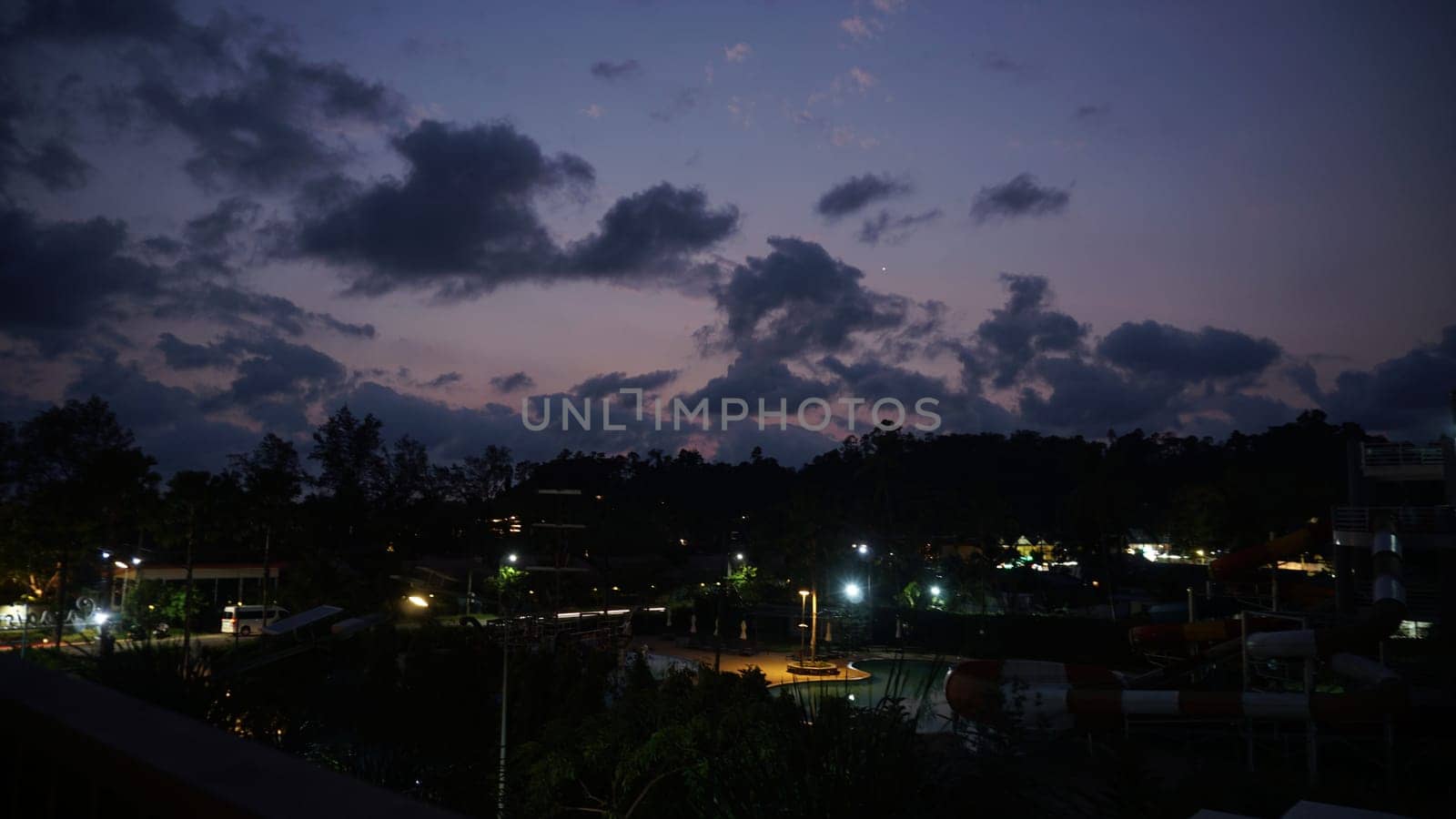 Streetlights casting a warm glow over a deserted water park at dusk, highlighting dramatic clouds and vibrant sunset colors painting the sky, creating a serene evening atmosphere