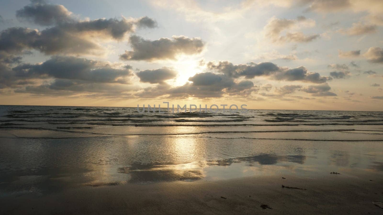 Soft sunlight filters through a cloudy sky, illuminating gentle waves rolling onto a wet sandy beach, creating a serene and reflective seascape at sunset