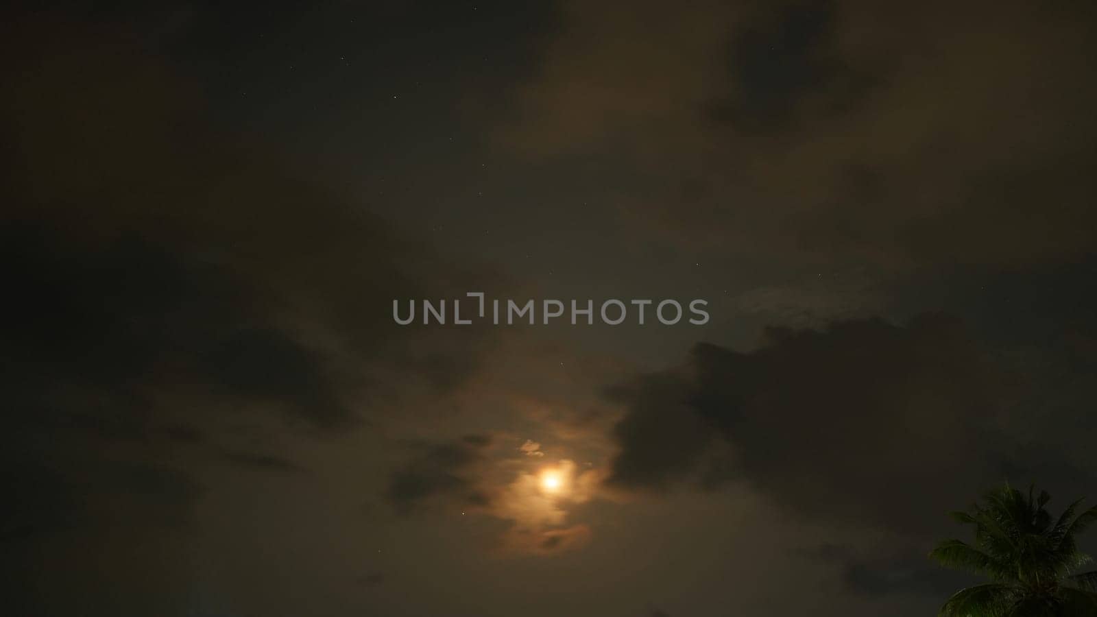 The moon shines through dark clouds at night, creating a moody and atmospheric scene with a few stars visible and a palm tree silhouette in the corner