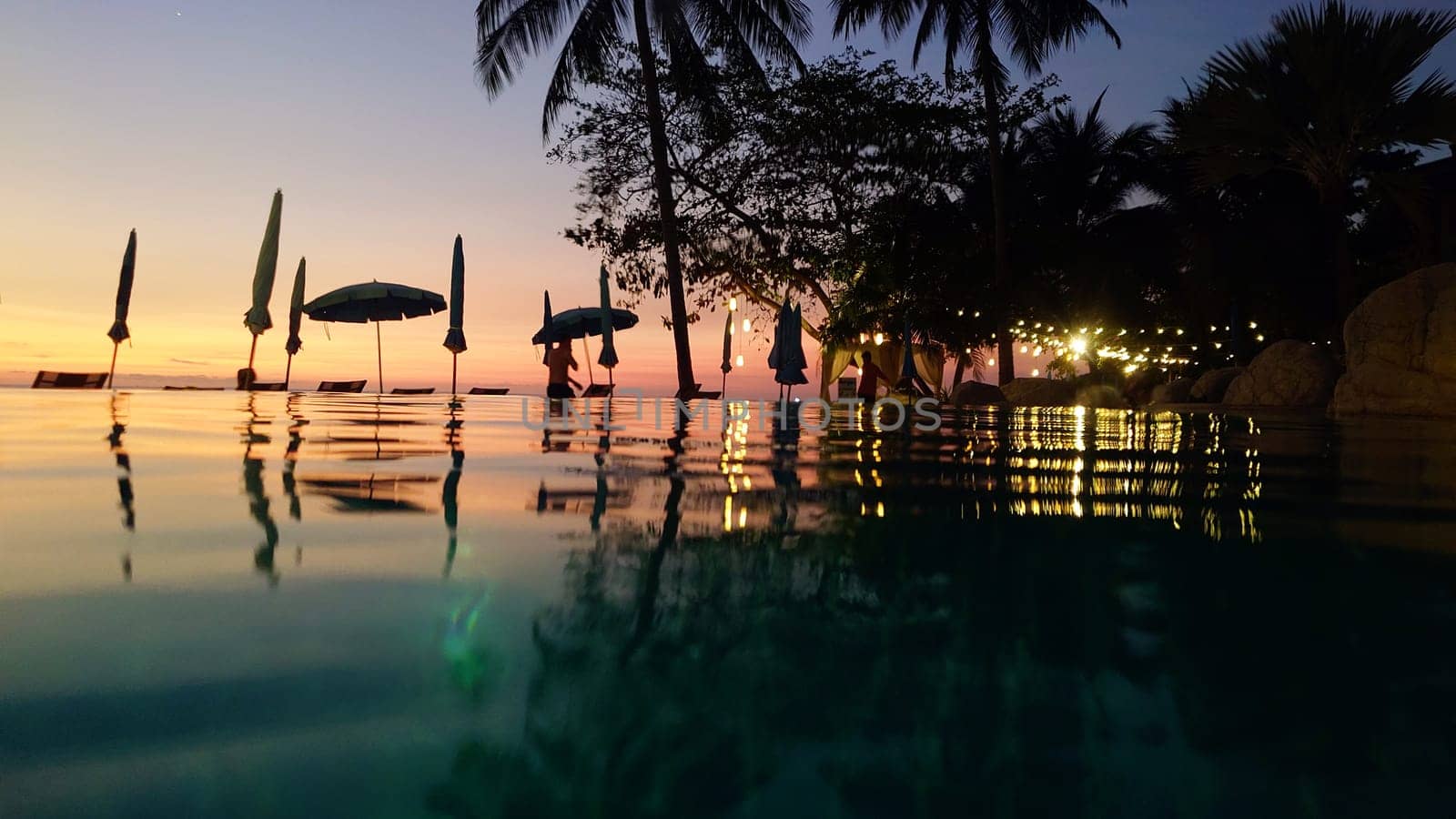 Tranquil infinity pool reflecting a beautiful sunset and string lights, creating a romantic atmosphere in a luxurious tropical resort with palm trees and parasols