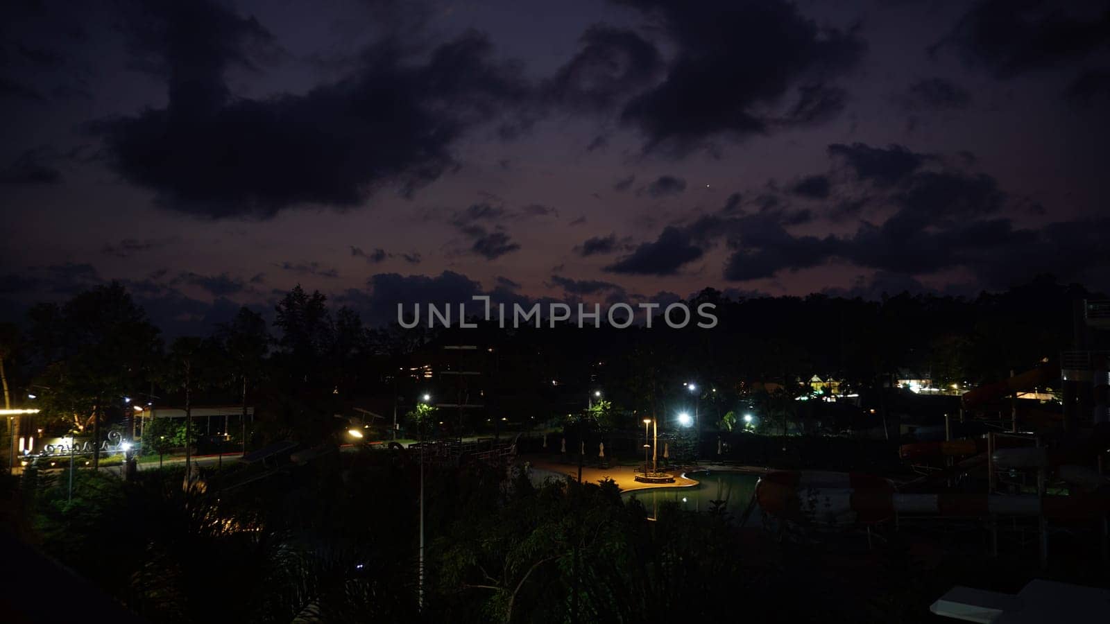 Streetlights casting a warm glow over a deserted water park at dusk, highlighting dramatic clouds and vibrant sunset colors painting the sky, creating a serene evening atmosphere