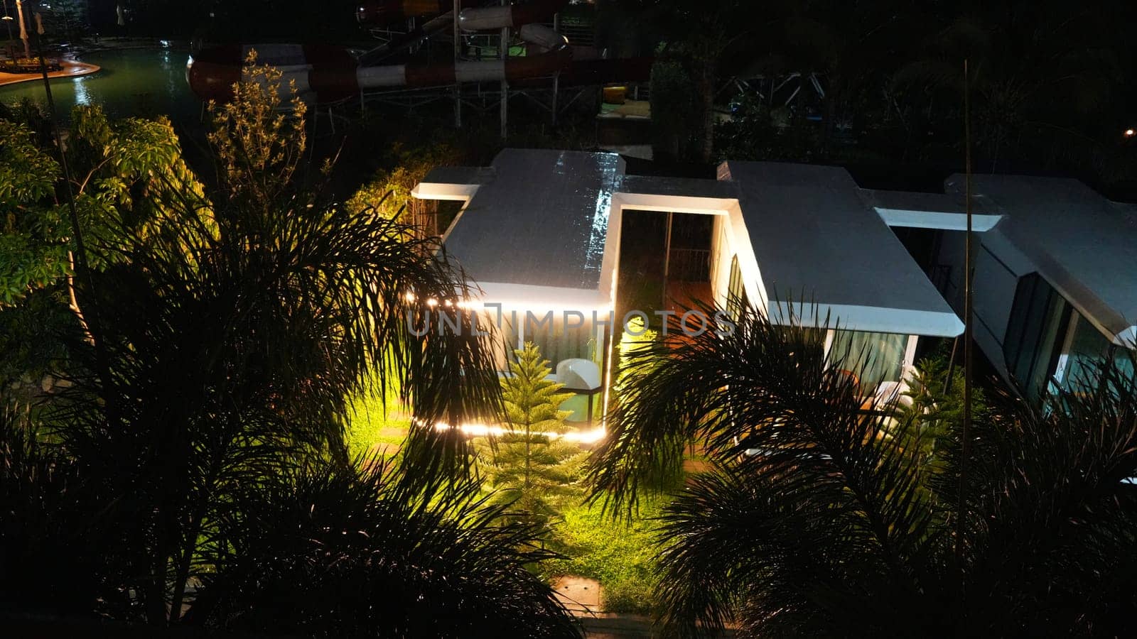 Warm lights illuminating modern bungalow at night, nestled among lush tropical foliage, with a water park shimmering in the background, creating a serene and inviting vacation atmosphere