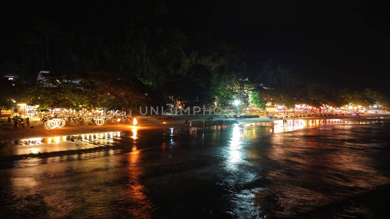 Tourists enjoying a vibrant fire dancing show on the beach, surrounded by restaurants at night, with colorful lights reflecting off the wet sand and shimmering sea