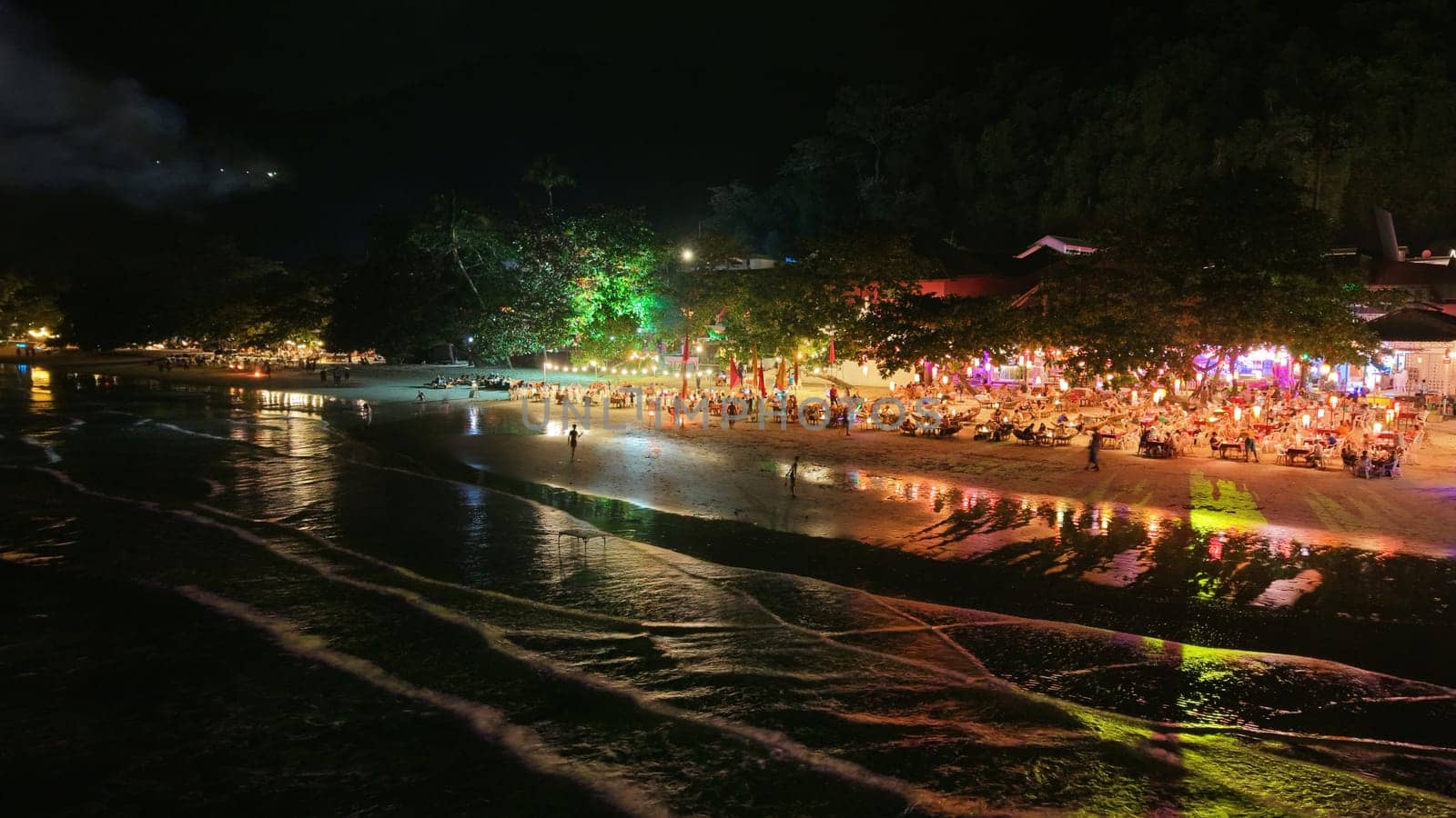 Tourists are dining and enjoying the vibrant atmosphere of a beachfront restaurant at night, with colorful lights reflecting on the wet sand and gentle waves lapping the shore