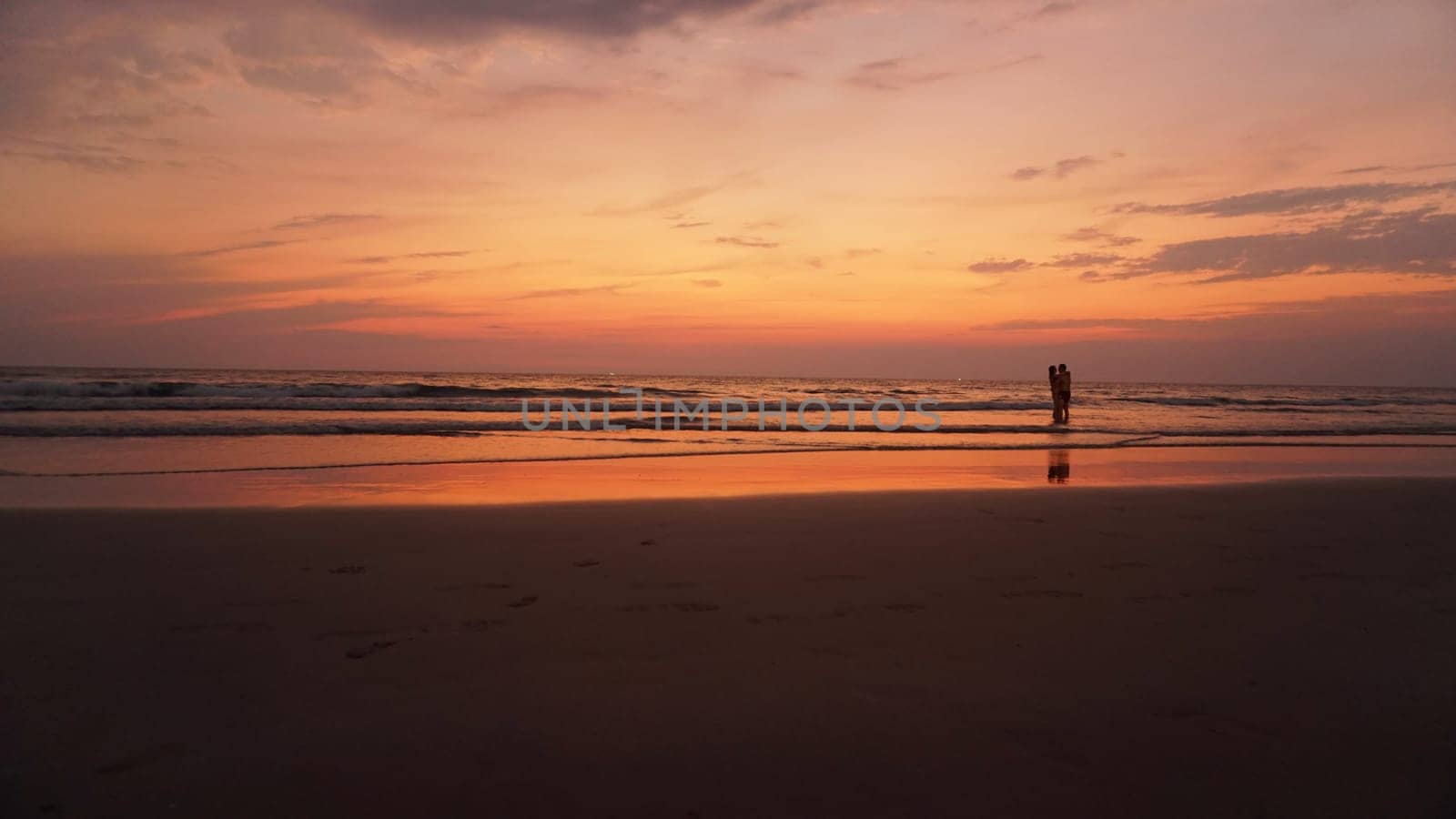 Golden hour paints the sky and ocean in warm hues as a couple embraces in the shallow water, creating a serene and romantic scene on a tropical beach
