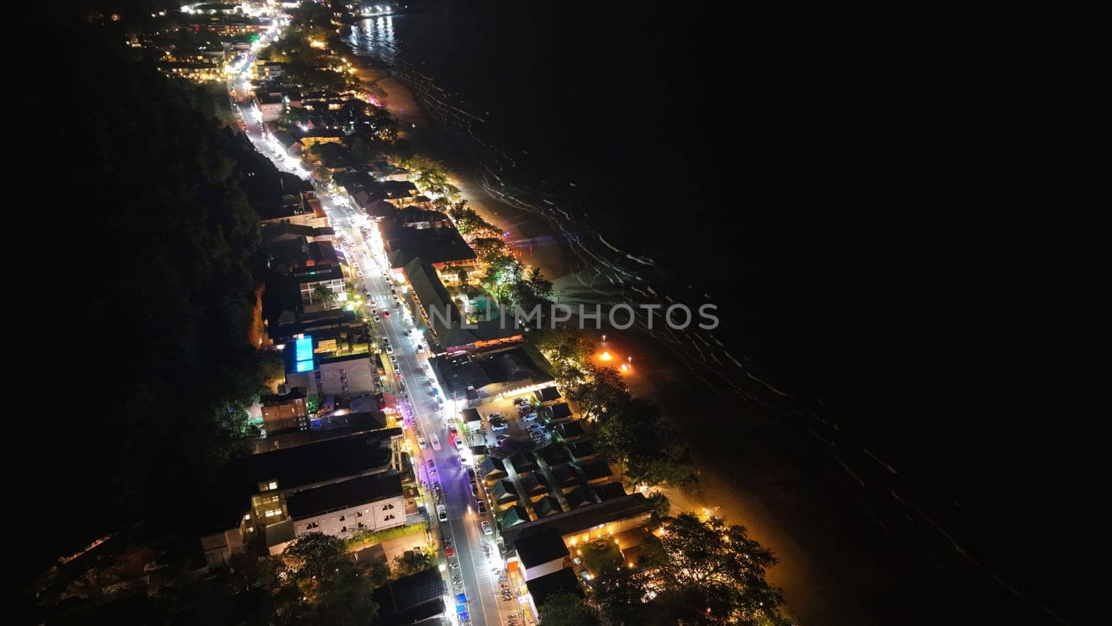 Capturing a vibrant coastal road at night, illuminated buildings, bustling traffic, and the dark ocean create a striking contrast between land and sea