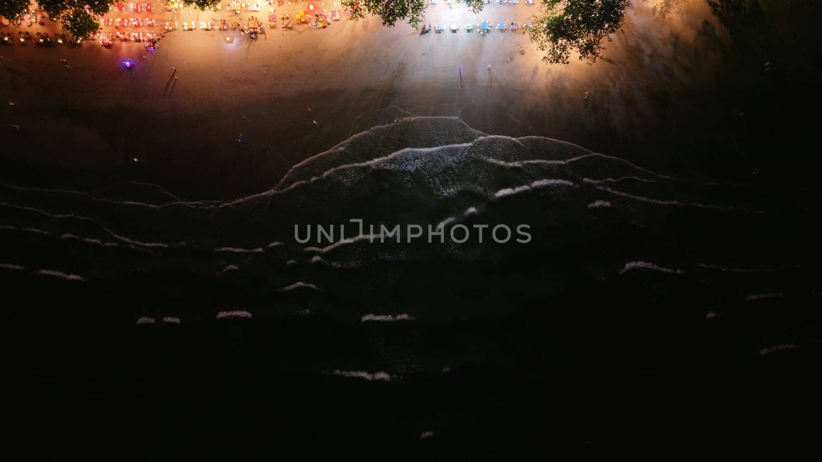 Gentle waves rolling onto a dark sandy beach at night, creating a serene atmosphere. Illuminated beach and trees in the background highlight a vibrant nightlife filled with joy and celebration