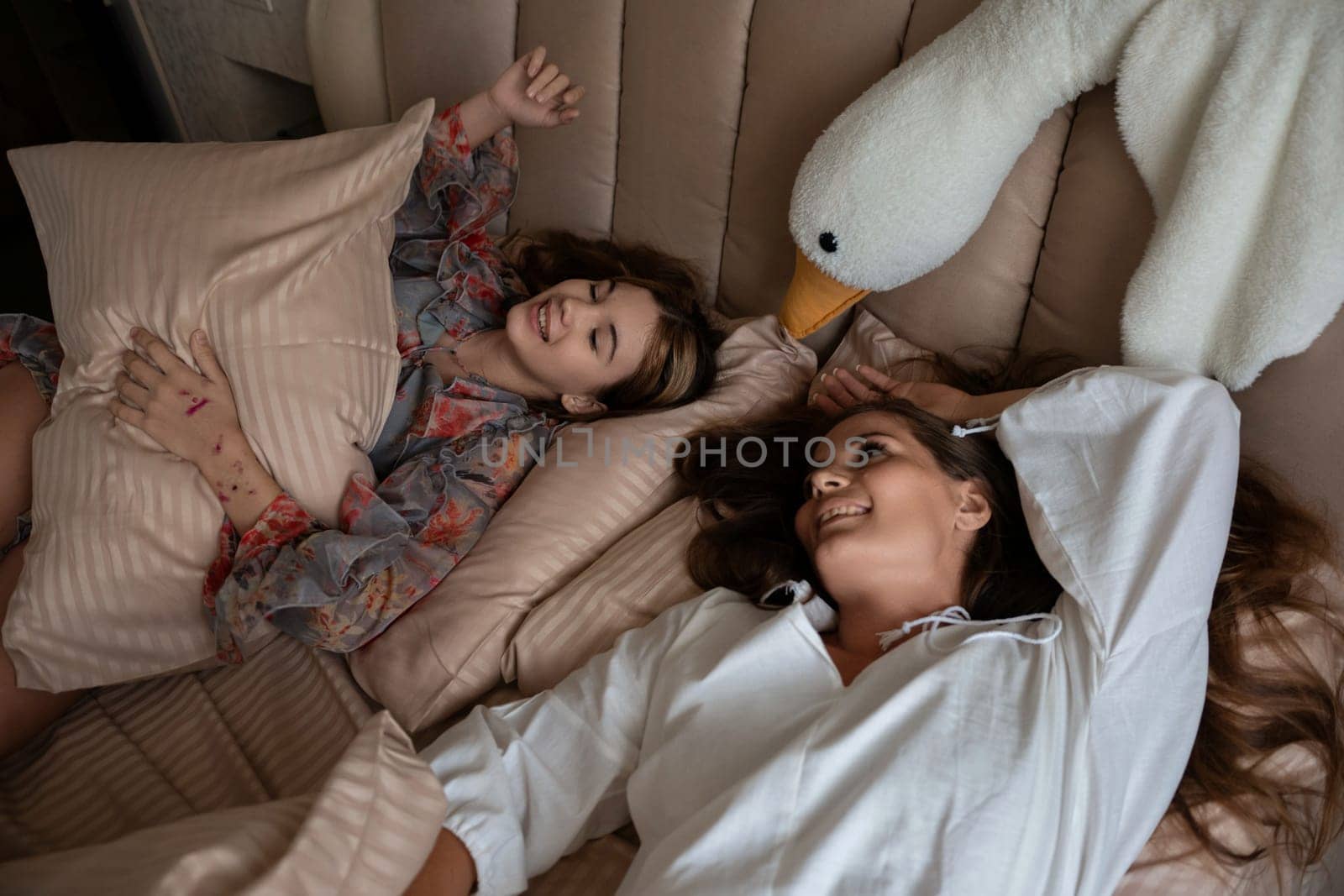 Two women lie in bed, smiling. One holds a pillow, the other rests, a large swan-shaped plush toy nearby.