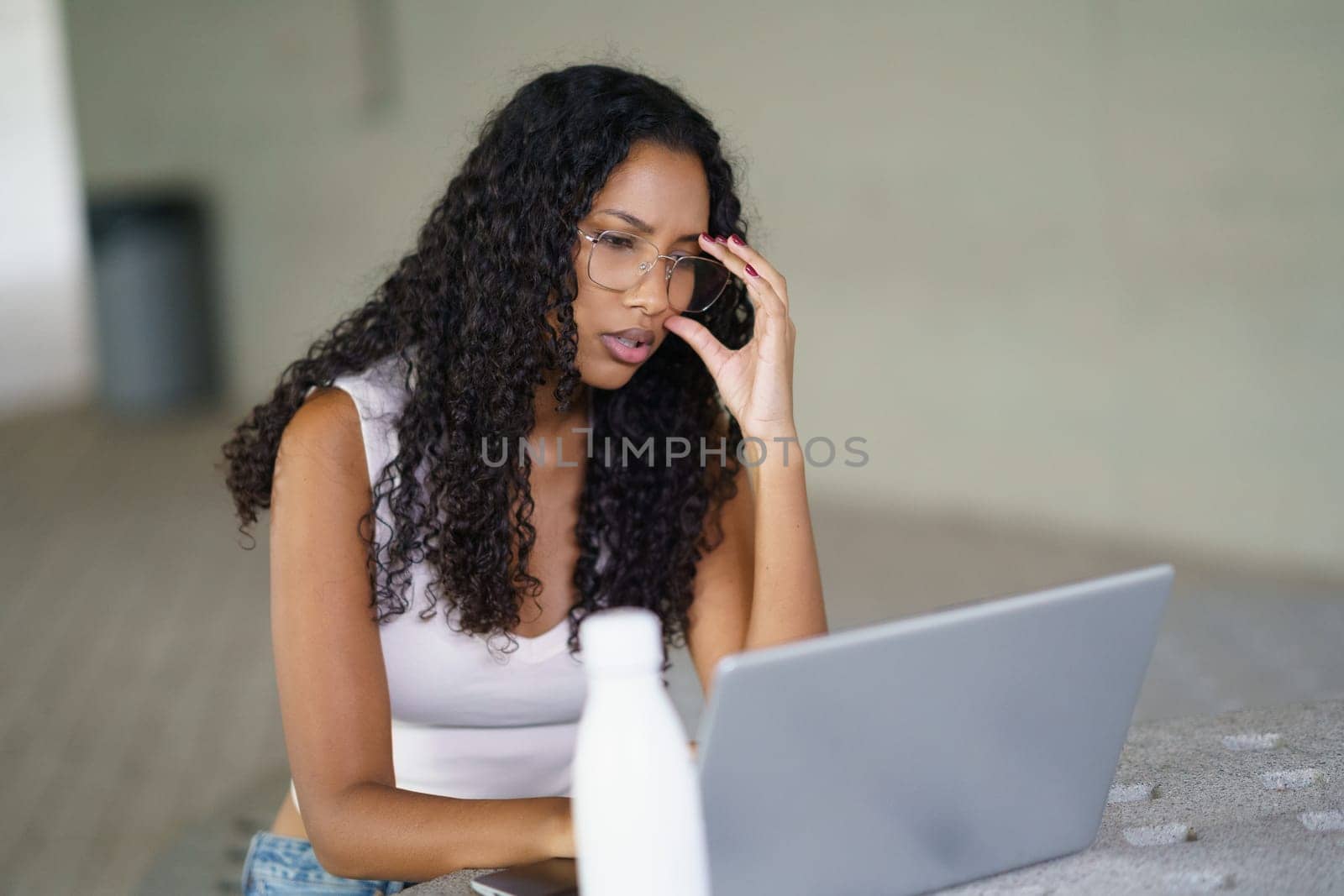 In a modern, contemporary environment, a woman with curly hair and stylish glasses is intensely concentrating on her laptop, showcasing her dedication and focus as she engages in task at hand