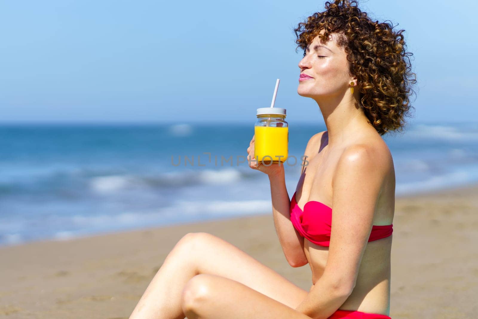 Calm curly female in bikini having glass cup of juice with straw while enjoying sunlight on sandy beach of ocean