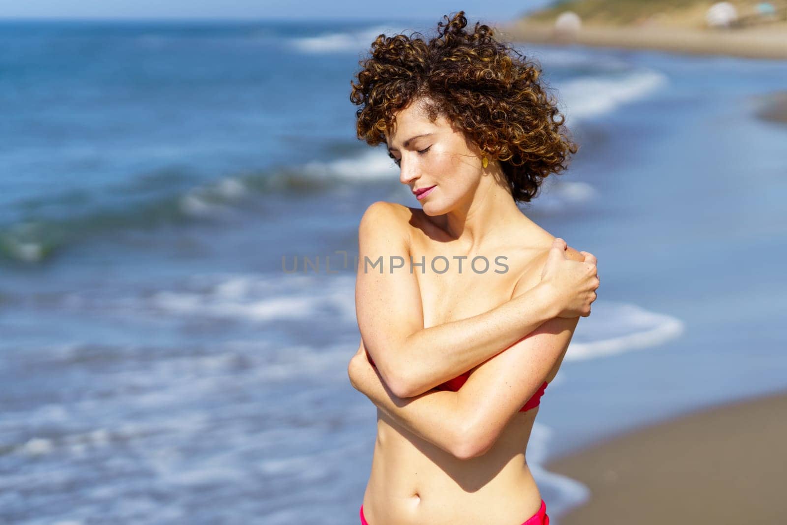 Smiling young female with ginger hair tilted head hands crossed and touching bare shoulder, while enjoying and standing in sunlight with eyes closed on blurred sea beach