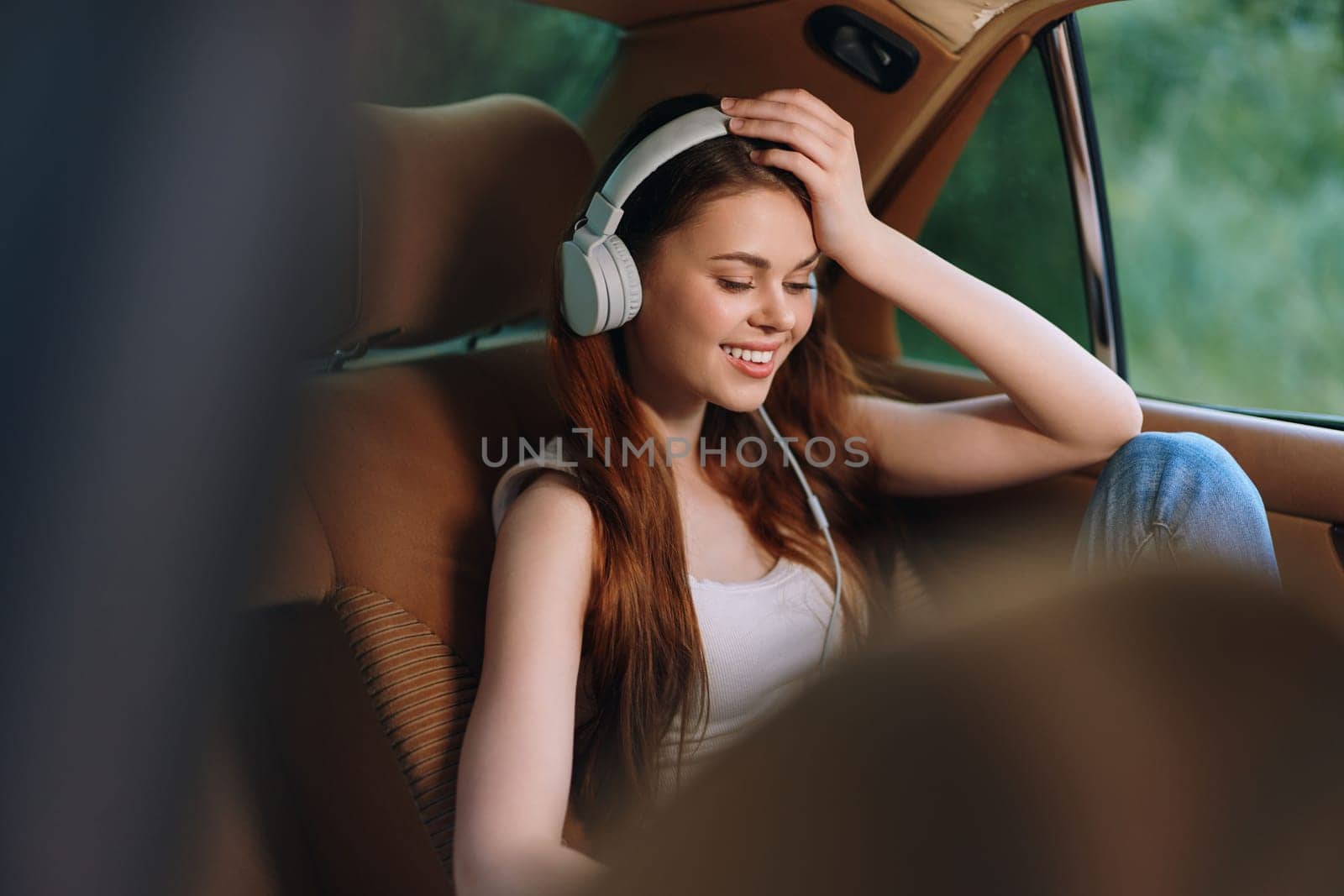 A cheerful young woman with long hair is sitting in a car, wearing headphones and smiling while listening to music. The soft, warm colors enhance a relaxed atmosphere, perfect for lifestyle and music