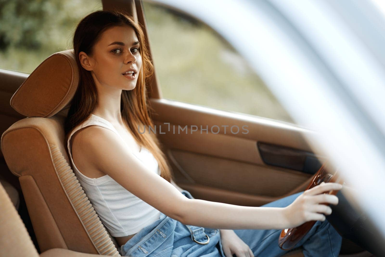 Confident young woman dressed in a simple white top and jeans, seated in a car's plush interior, capturing the essence of summer vibes and freedom on the road.