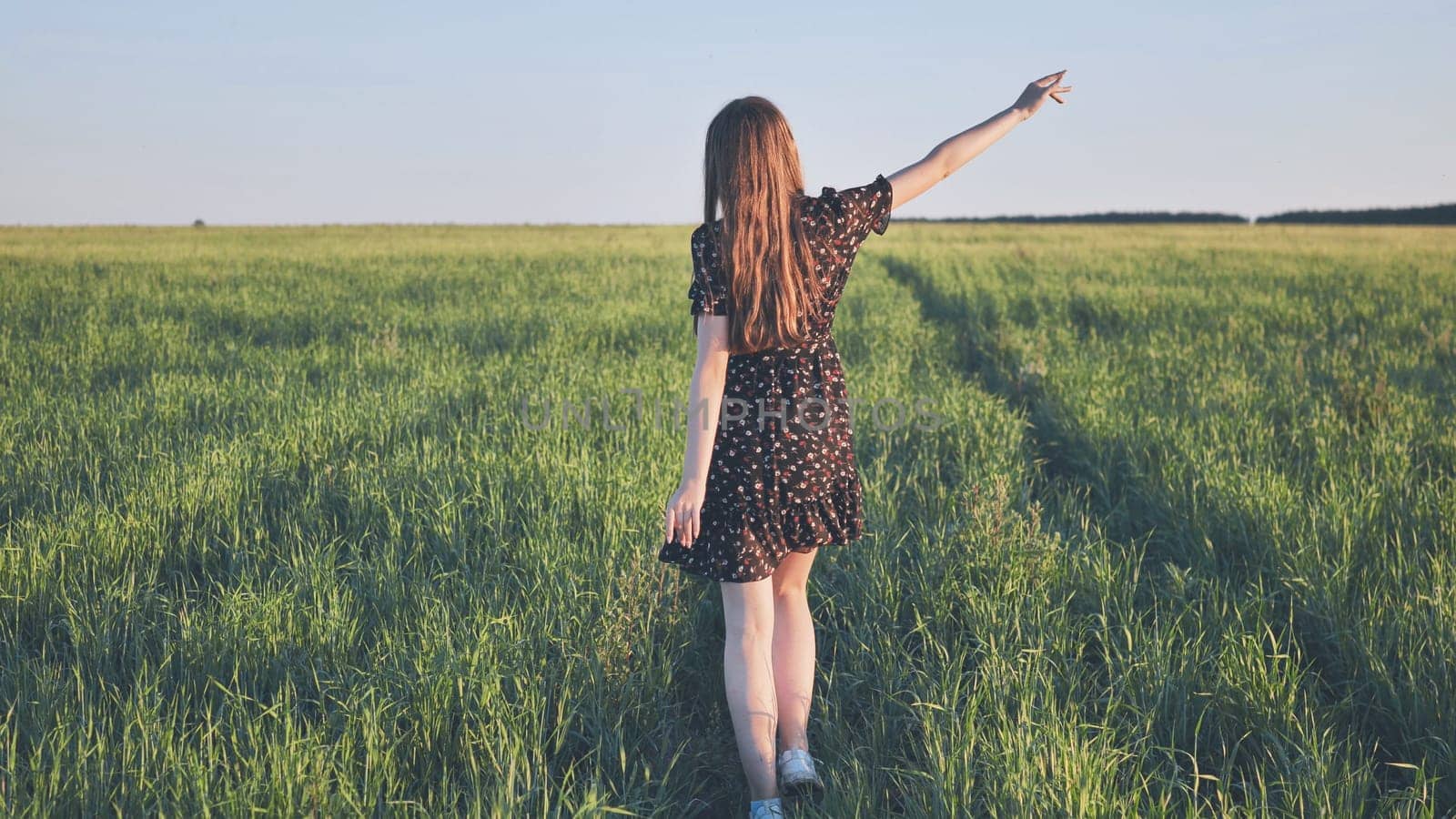 Young woman with long brown hair walks through a green field at sunset, her arm outstretched, enjoying the peace and beauty of nature