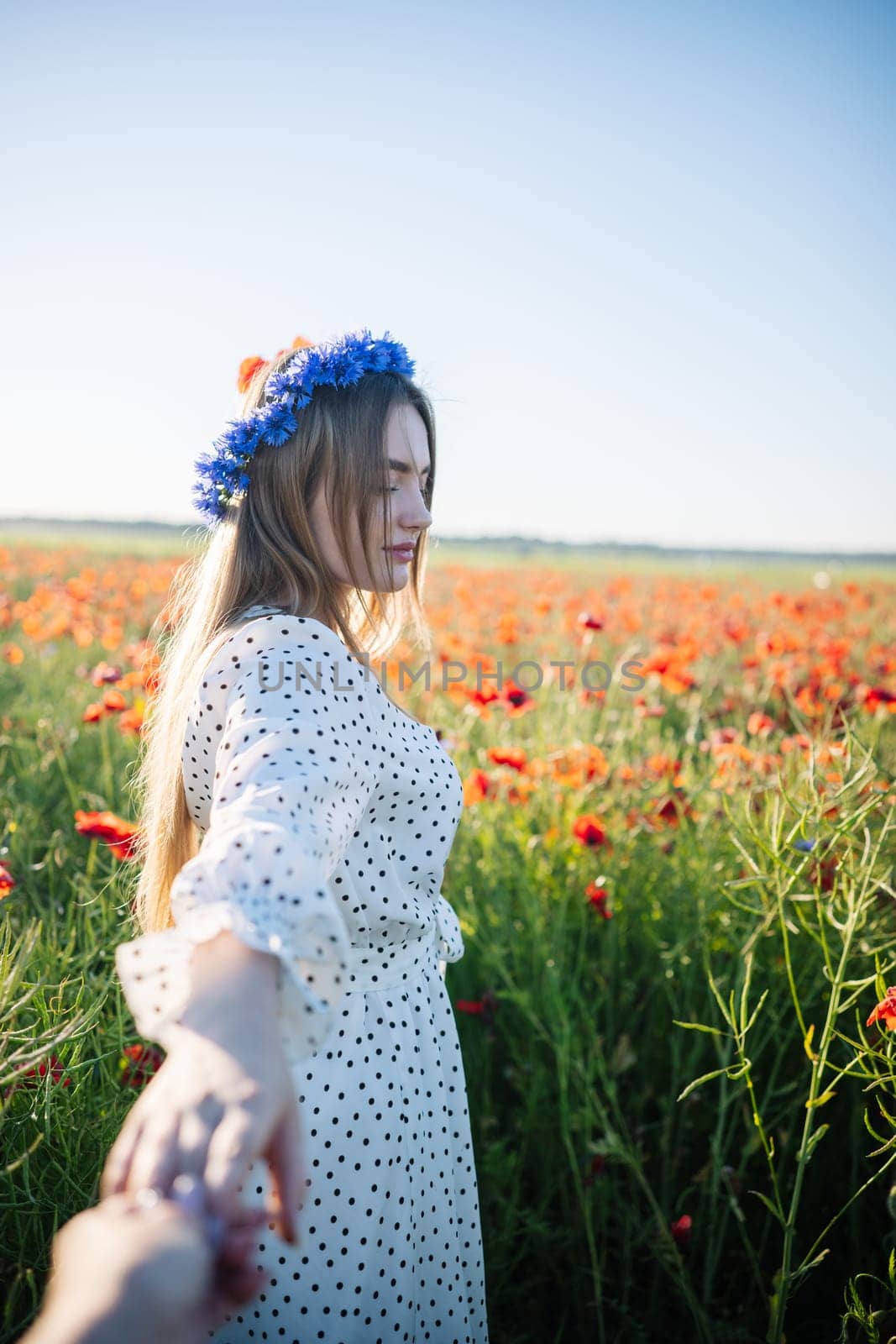 Woman with cornflower wreath walking through blooming poppy field, holding invisible partner's hand
