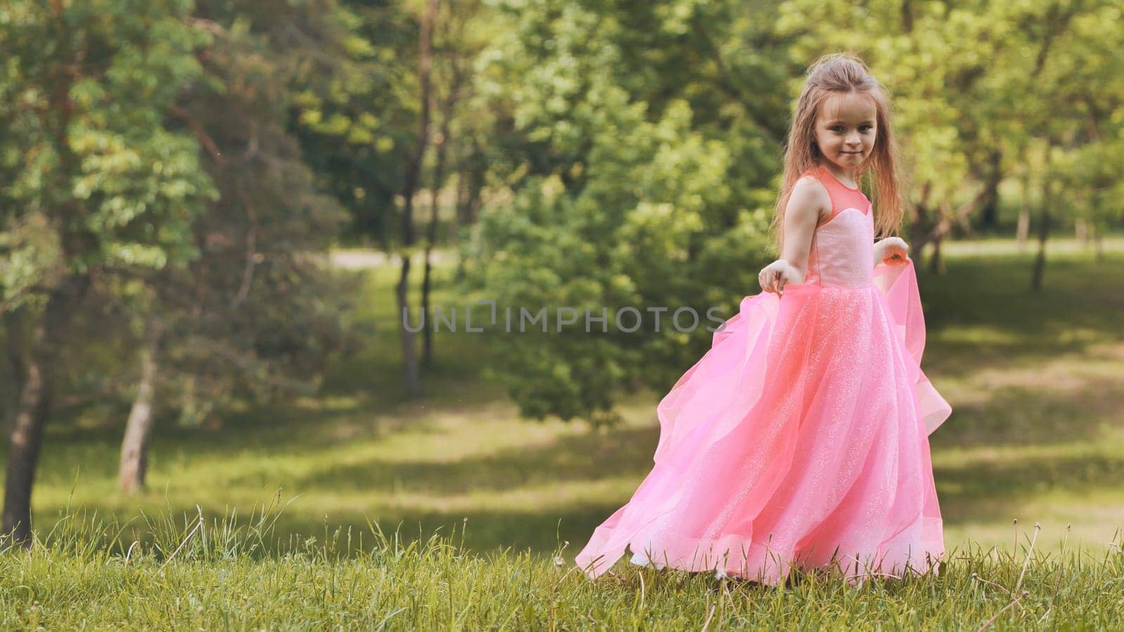 Cute child wearing beautiful pink dress walking in a green park during a sunny summer day