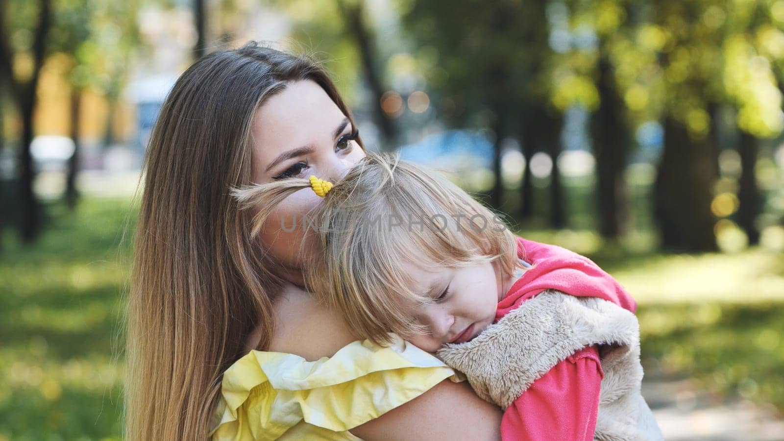 Caring mother hugging tearful daughter near trees, offering emotional support during gentle sunlit afternoon in verdant park setting