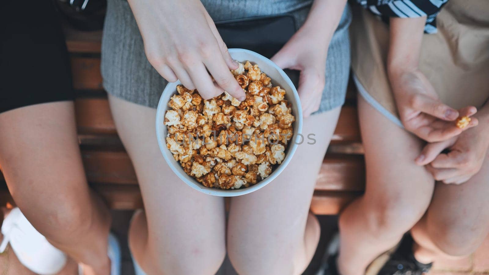 Young woman sharing caramel popcorn with friends, laughing and relaxing on sunlit park bench during casual afternoon gathering
